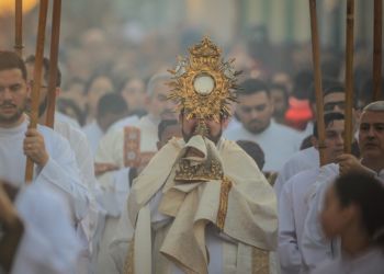 Padre carrega o Corpo de Cristo. Multidão acompanha a procissão